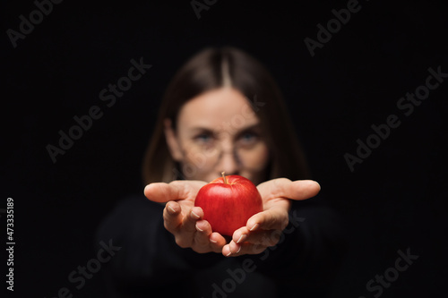 A smiling woman wearing glasses holding a red apple in palms show it to camera, with red lips, over a black background. Focus on fruit