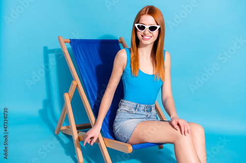 Portrait of attractive cheerful girl sitting in chair sun bath spending free time isolated over bright blue color background #473341730