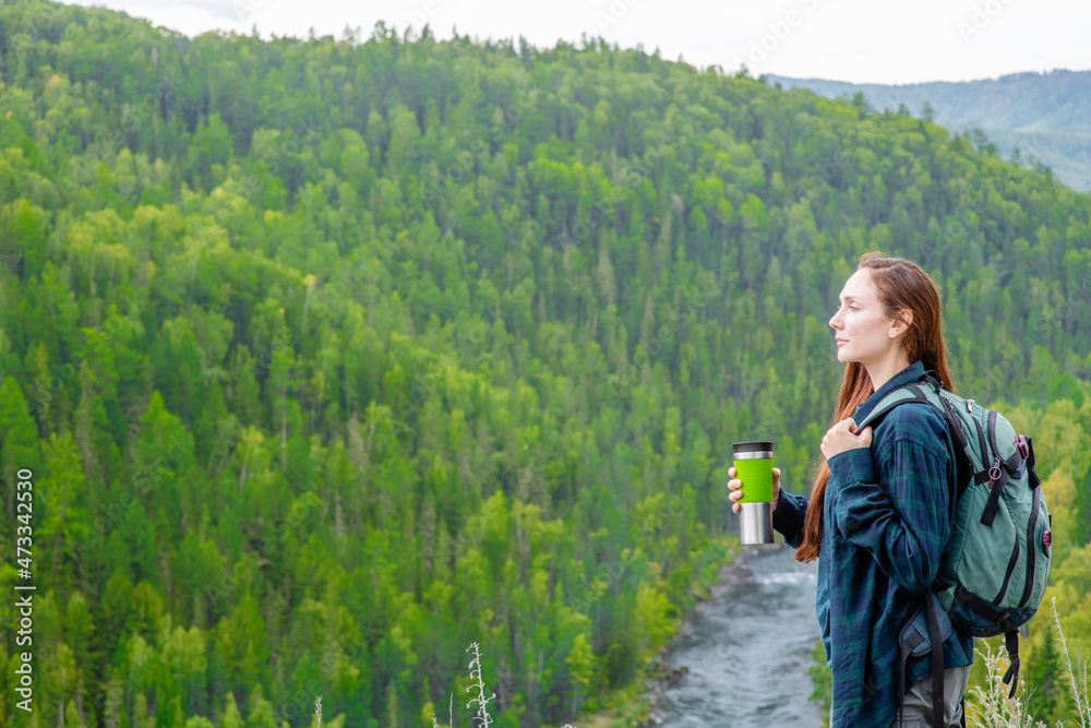 Young woman with backpack and thermos enjoys the view of nature at hike. Empty space for text