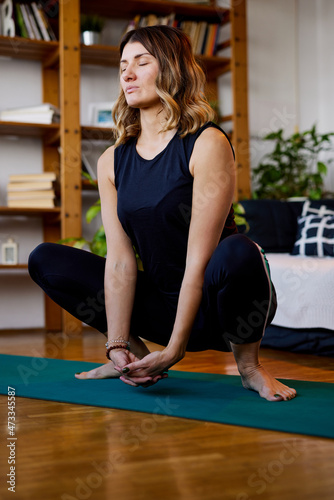 A middle-aged yogi woman crouches in Garland yoga pose and meditates with eyes closed. A yogi woman relaxing at home.