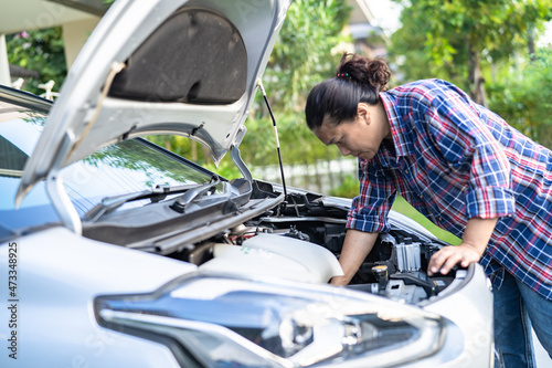 Asian woman looking and check under the hood of broken down car on the side of the road.