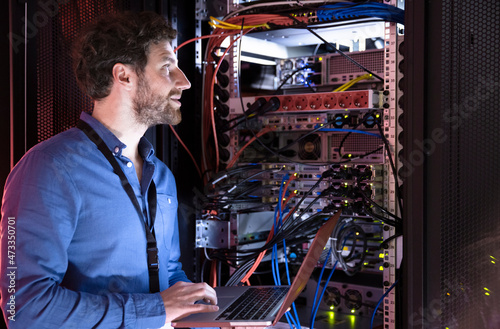 Male IT professional looking away while holding laptop by server rack in data center photo