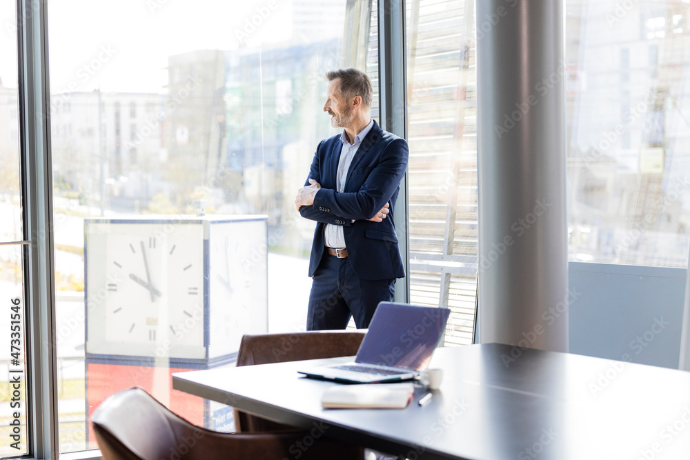 Businessman with arms crossed looking through window in office Businessman with arms crossed looking through window in office
