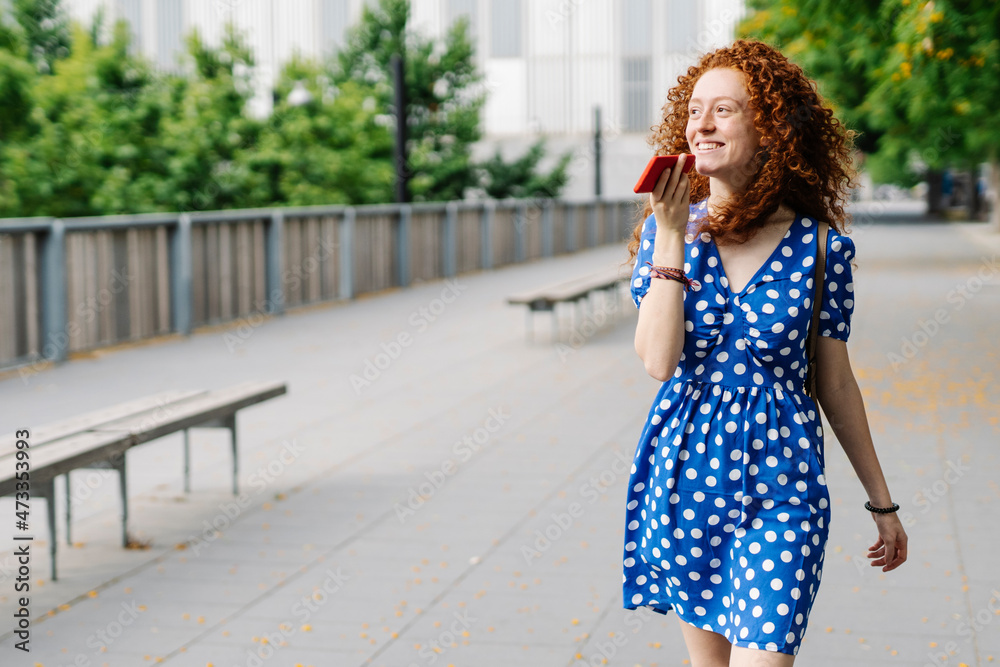 Smiling woman sending voicemail through mobile phone while walking on footpath Smiling woman sending voicemail through mobile phone while walking on footpath