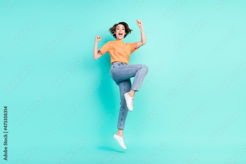 Photo of cute lucky young lady wear orange t-shirt jumping high rising fists smiling isolated turquoise color background