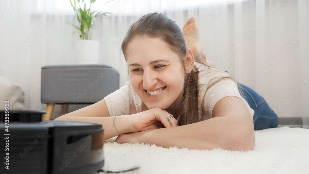 Portrait of happy smiling woman lying on carpet and looking on working robot vacuum cleaner. Concept of hygiene, household gadgets and robots at modern life. Portrait of happy smiling woman lying on carpet and looking on working robot vacuum cleaner. Concept of hygiene, household gadgets and robots at modern life.