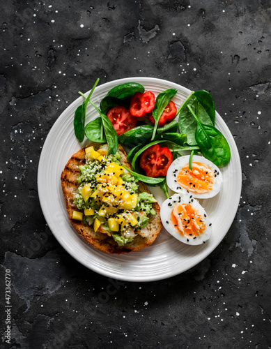 Delicious healthy breakfast, snack - sandwich with avocado, shrimp, mango salsa, boiled egg and spinach tomatoes salad on a dark background, top view