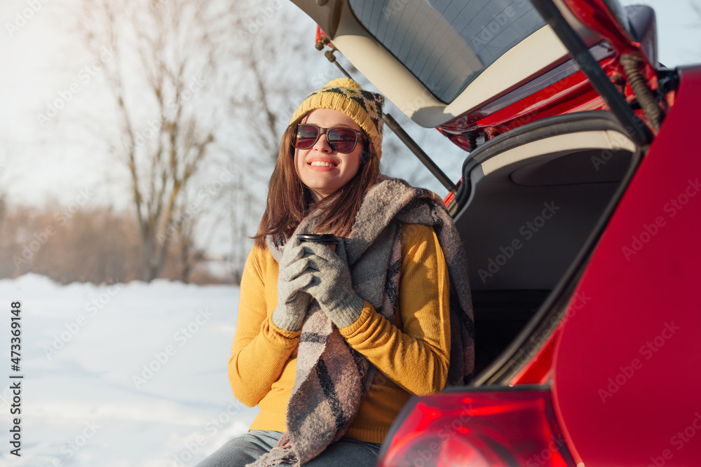 Woman with coffee in hands is sitting in car trunk and has winter picnic
