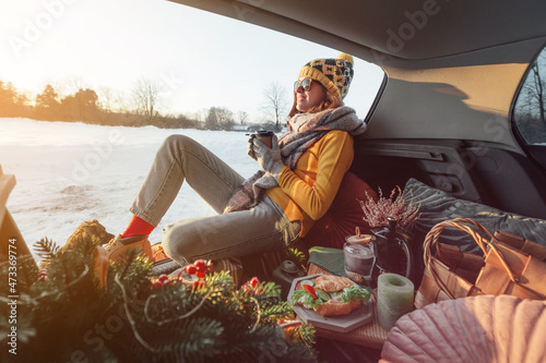 Woman with coffee in hands is sitting in car trunk and has winter picnic