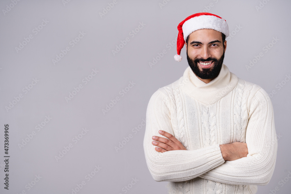 Smiling handsome middle-eastern man in Santa Claus red hat looking at camera, preparing for Christmas New Year celebration isolated over grey background