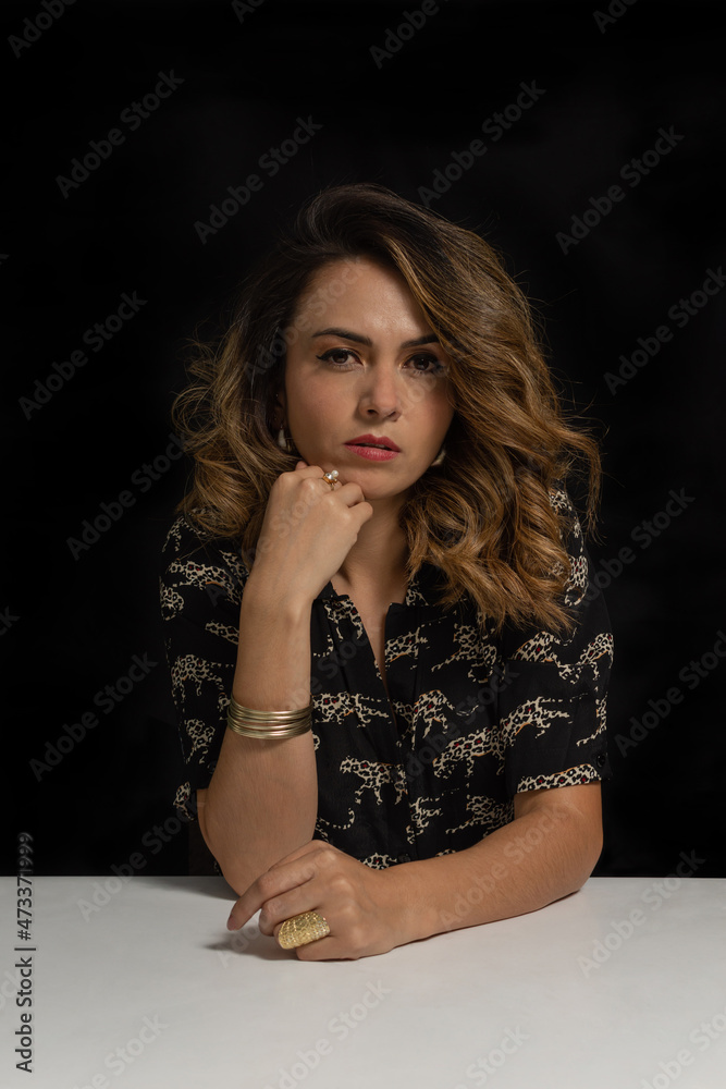Latin young woman sitting with serious expression isolated on black background Latin young woman sitting with serious expression isolated on black background