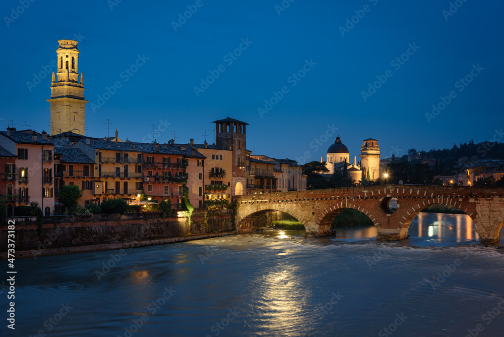 City landscape with the old town buildings along the bank of the Adige river and the famous Stone Bridge (Ponte di Piettra) at night, Verona, Veneto Region, Italy