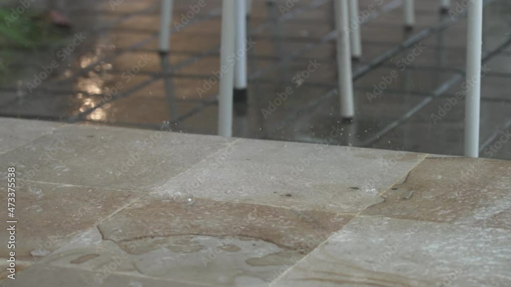 Close-up of wet marble floor tiles of the restaurant terrace with white chairs in heavy rain. Puddles with streaks from heavy rain in slow motion. A modern restaurant on a cloudy day.