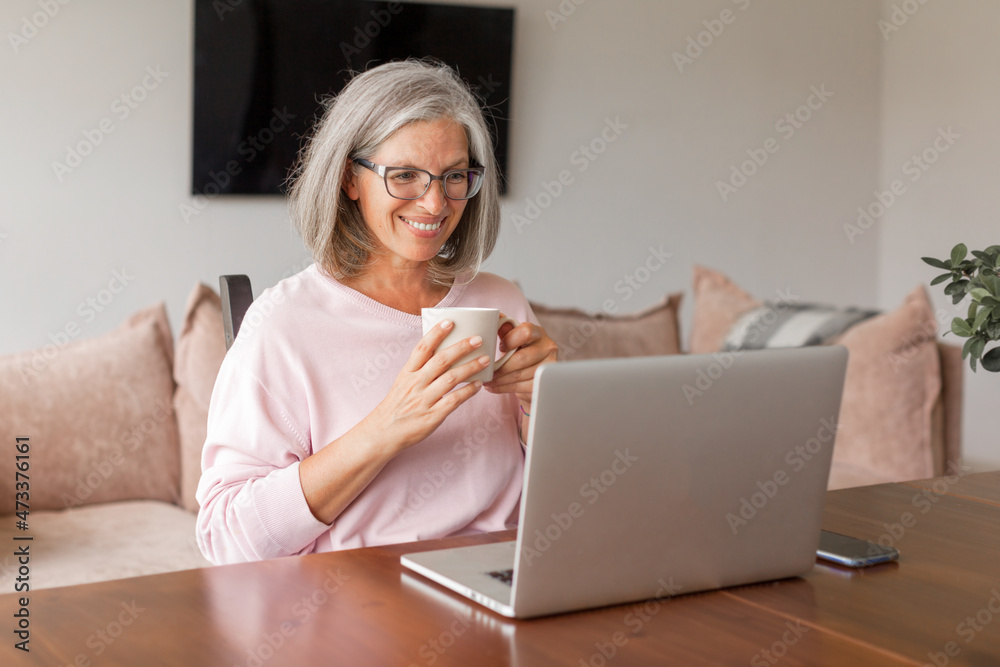 Happy middle age senior woman sitting at the table at home working using computer laptop Happy middle age senior woman sitting at the table at home working using computer laptop
