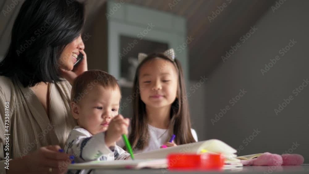 Happy Asian mom talking by phone and looking after children in the kitchen Happy Asian mom talking by phone and looking after children in the kitchen