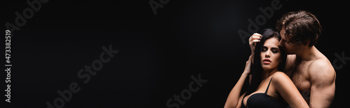 passionate man smelling hair of brunette woman in bra isolated on black, banner.
