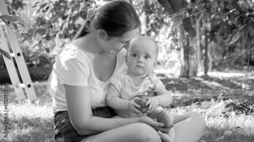 Black and white portrait of little baby boy holding apple sitting with mother on grass at park