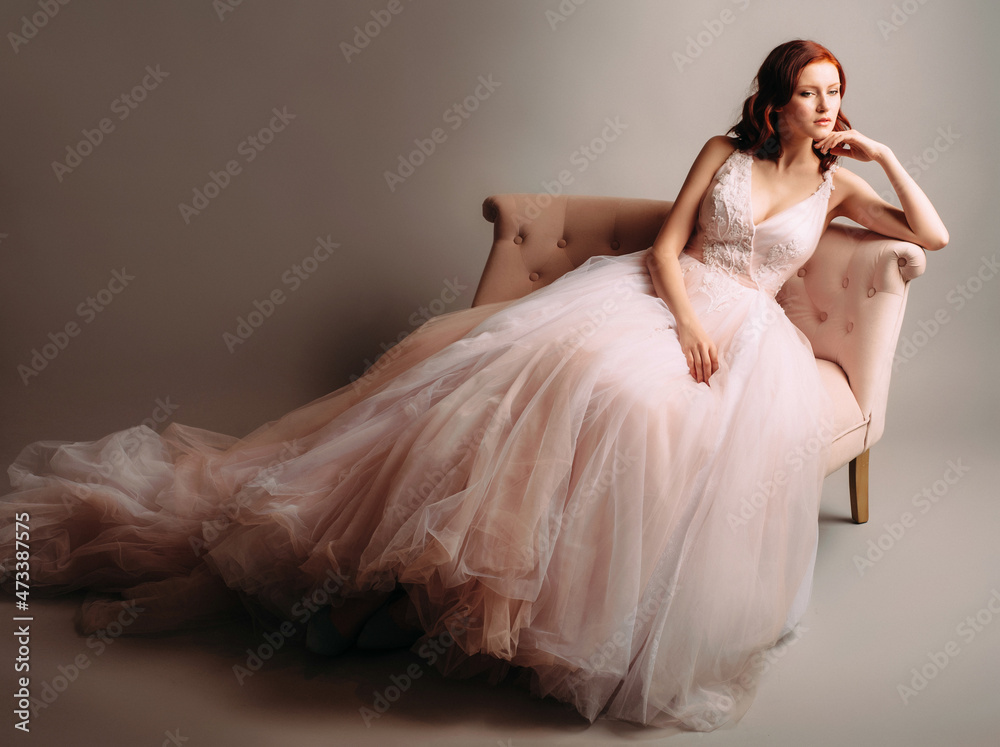 Fototapeta premium Redheaded bride in a pink wedding dress sitting in a vintage sofa. Studio portrait on neutral background.