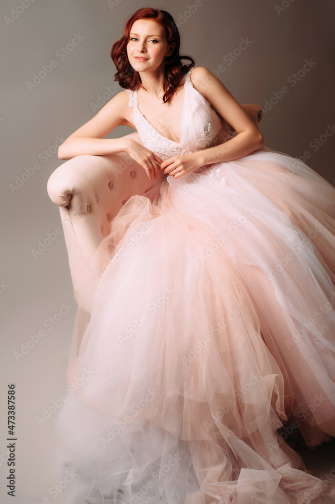 Fototapeta premium Happy ginger girl in pink wedding dress on grey background. Gorgeous bride on pale pink armchair in neutral studio room. Fashion shooting.