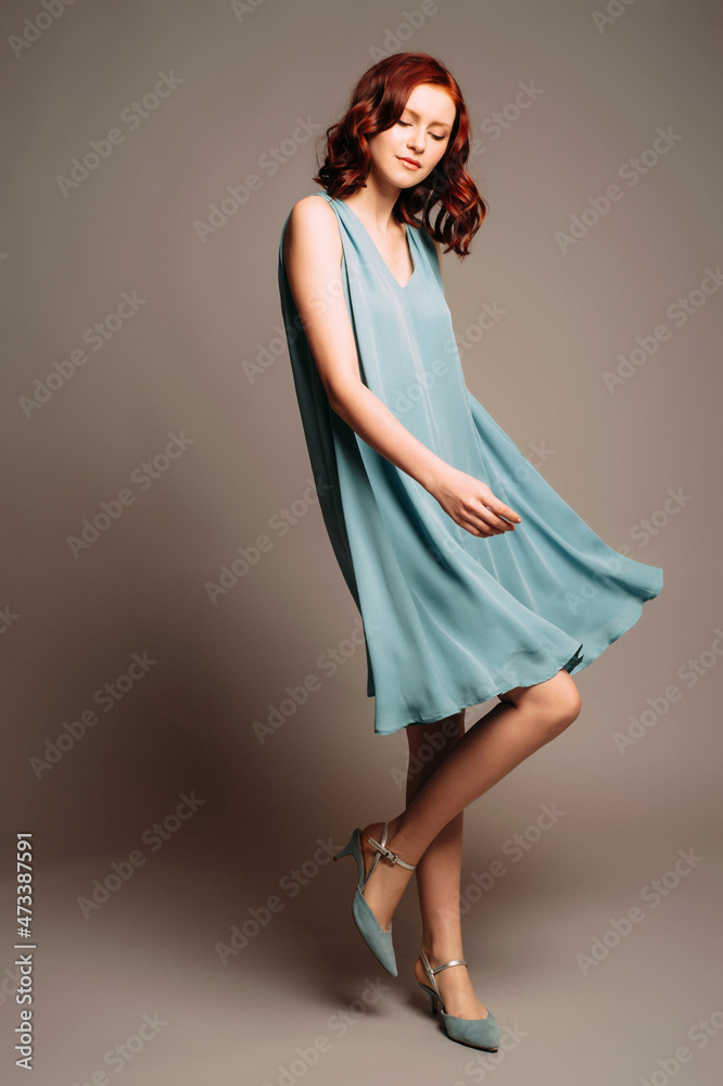 Fashion look in dust blue. Beautiful portrait of ginger young woman in summer fluttering dress and suede shoes on grey background at studio.