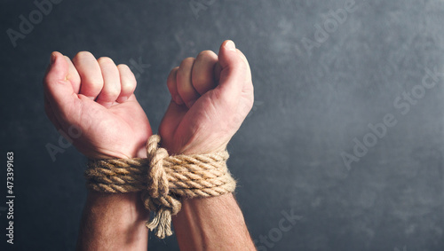 Male hands tied with a rough rope at the wrists close-up on a dark background, soft focus. Conceptual image of violence, slavery, dependence, lawlessness, lack of freedom photo