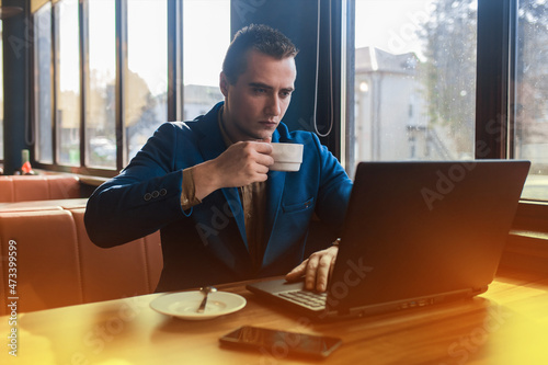 A business busy man businessman a stylish portrait of Caucasian appearance in a jacket, works in a laptop or computer, drink coffee on coffee break, sitting at a table by the window in a cafe