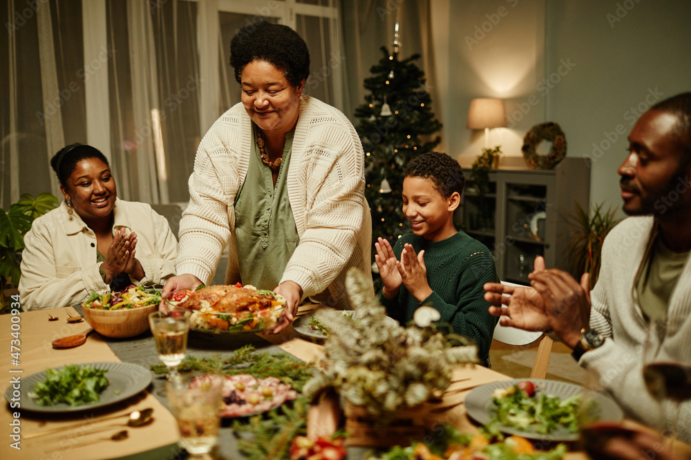 Portrait of smiling African-American grandmother bringing food to table while celebrating Thanksgiving with big happy family