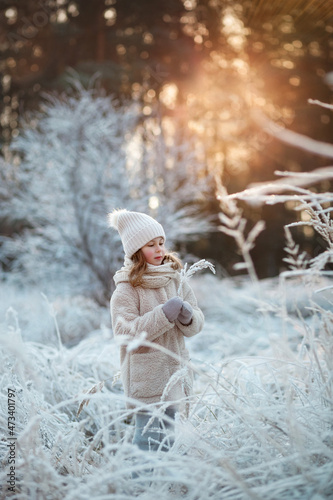 Frosty sunny winter day child girl in a fur coat and knitted hat holding a branch covered with hoarfrost