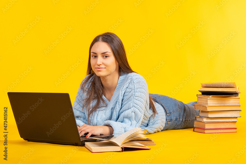 Woman lies with laptop and books. Joyful woman with textbooks. Woman student is engaged in self-education. University student does her own work. Stack of books near student. College girl on yellow Woman lies with laptop and books. Joyful woman with textbooks. Woman student is engaged in self-education. University student does her own work. Stack of books near student. College girl on yellow