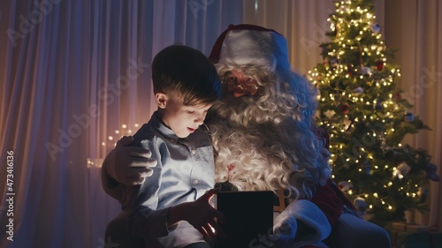 Little cute boy sitting on Santa's knees, opening a gift in front of Christmas tree. Adorable child is happy and surprised.