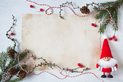 Christmas white wooden background with old paper for congratulation text, list, letter to Santa and fir branches, toy Santa, cones, red berries, red and white rope.