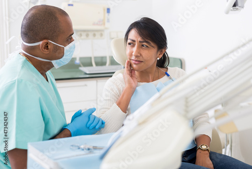 Latina suffering from toothache talking to stomatologist while sitting on dental chair in clinic