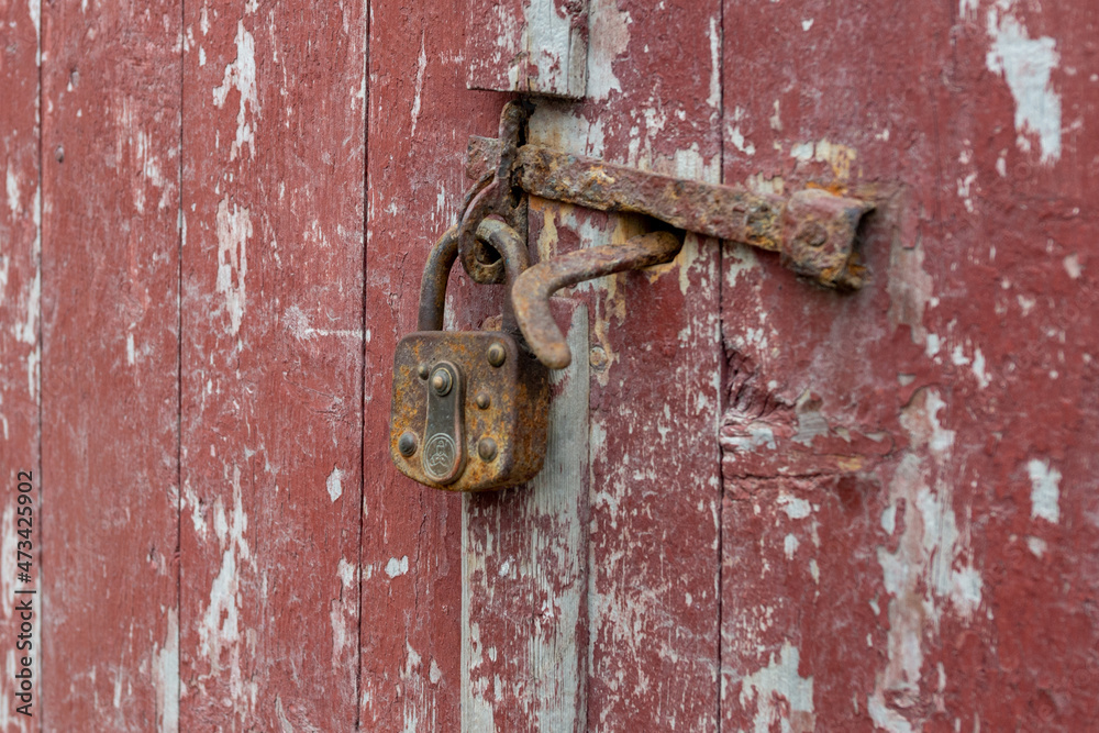 An antique rusty padlock attached to an old metal latch on a red wooden panel door. The building trim is white wood with a line pattern. The lock has a keyhole cover and the latch is black metal. An antique rusty padlock attached to an old metal latch on a red wooden panel door. The building trim is white wood with a line pattern. The lock has a keyhole cover and the latch is black metal.