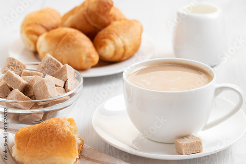 Coffee with milk in a white cup, buns and brown sugar on a white wooden table