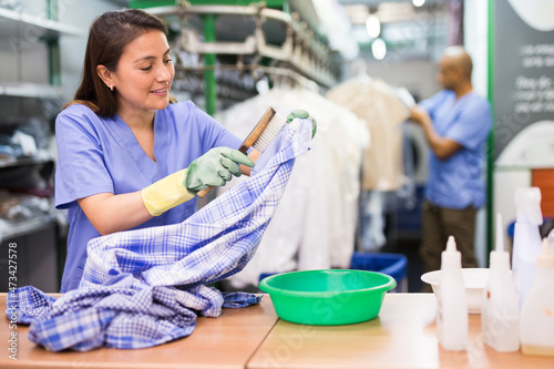 Focused female drycleaner removing spots and stains cleaning shirt using chemicals and brush