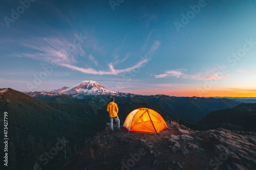 A man is standing by a tent on the top of a mountain near mt. Rainier