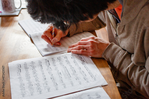 A man sits at a wood table transcribing sheet music with a pencil photo