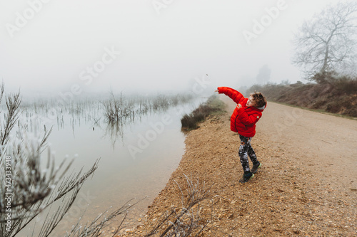 Boy in red coat throwing stone into lake against foggy overcast sky photo