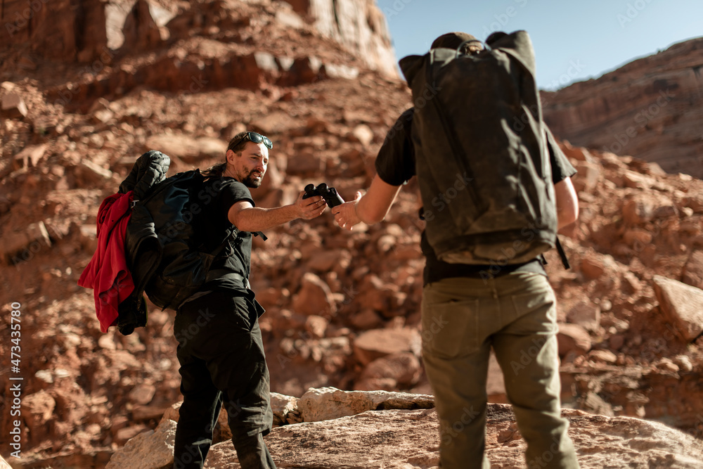 Man giving binoculars to friend while hiking on mountain at Canyonlands National Park Man giving binoculars to friend while hiking on mountain at Canyonlands National Park