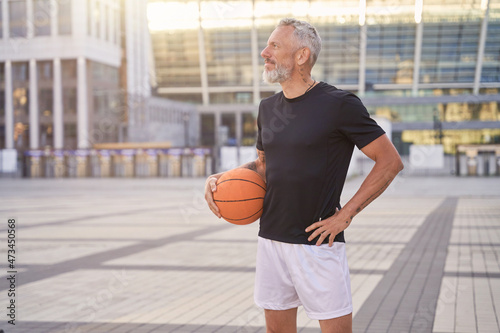 Thoughtful sportive mature man in sportswear standing outdoors with basketball ball, ready for workout © Kostiantyn