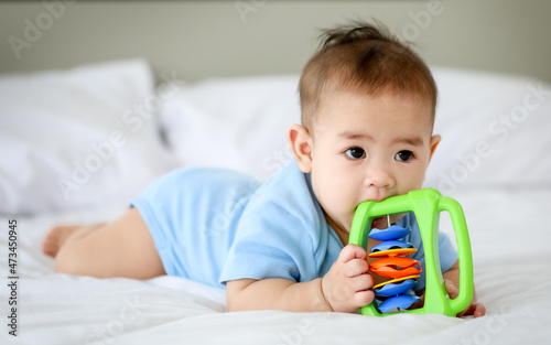 Adorable and innocent little Asian baby enjoys playing fun with colorful children toy while lying on stomach on bed.