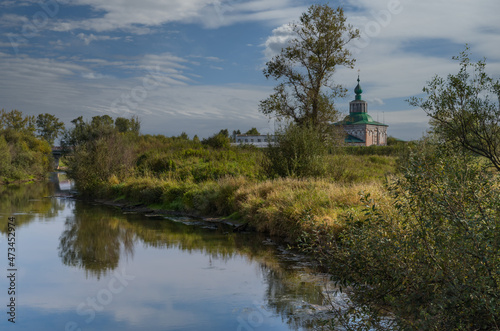 Summer landscape in the city limits of Solikamsk (Russia). The raised bank of the river, overgrown with grass, trees with kutarniks, which are reflected in the water. In the distance the old church