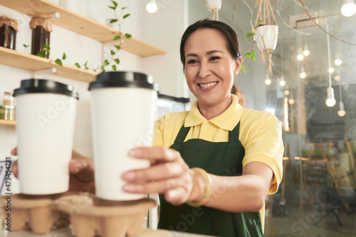 Smiling mature coffeeshop worker giving customer two cups of take out coffee