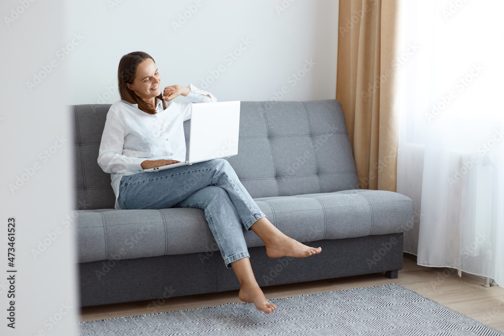Smiling positive Caucasian female freelancer wearing white shirt and jeans sitting on cough and working on notebook, looking at pc screen and expressing happiness, having pleasant online conversation. Smiling positive Caucasian female freelancer wearing white shirt and jeans sitting on cough and working on notebook, looking at pc screen and expressing happiness, having pleasant online conversation.