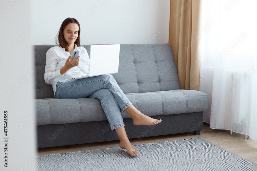 Smiling positive young adult woman wearing white shirt and jeans sitting on sofa at home and working on laptop and browsing Internet via smart phone while having break. Smiling positive young adult woman wearing white shirt and jeans sitting on sofa at home and working on laptop and browsing Internet via smart phone while having break.