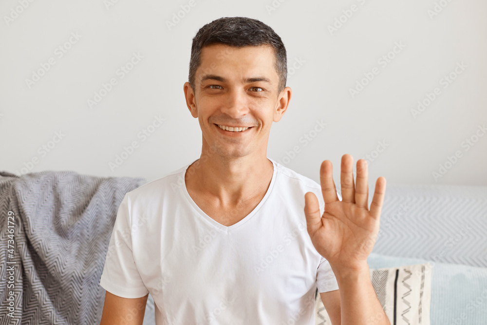 Portrait of happy optimistic dark haired young adult male wearing white casual style t shirt sitting on sofa, looking at camera and waving hand, saying hello or bye. Portrait of happy optimistic dark haired young adult male wearing white casual style t shirt sitting on sofa, looking at camera and waving hand, saying hello or bye.