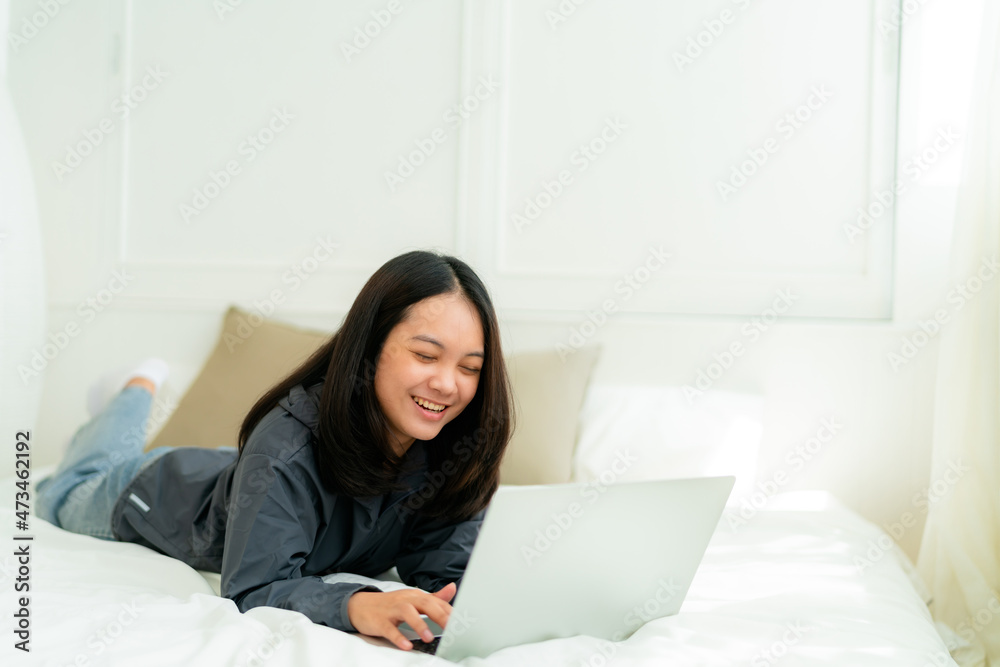 Portrait, a cute Asian teenage girl. wearing gray shirt lying in bed with a notebook computer in the bedroom to study online with a happy face Portrait, a cute Asian teenage girl. wearing gray shirt lying in bed with a notebook computer in the bedroom to study online with a happy face
