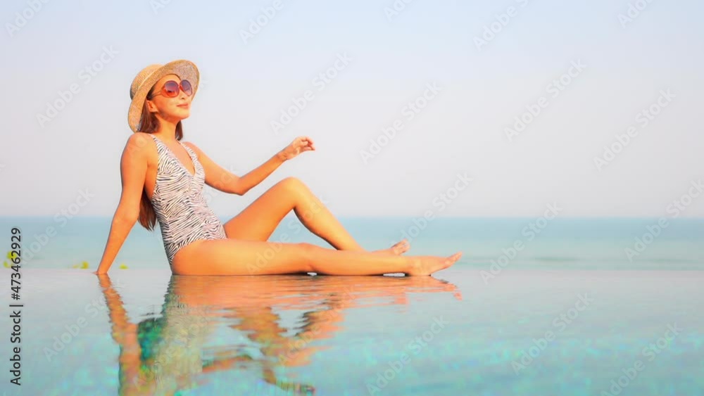 Asian Woman in Blotchy Swimming Suit Sitting on the Edge of Infinity Pool Touching the Frame of Red Sunglasses on Turquoise Seascape Background on a Sunny Day Slow-Motion side view