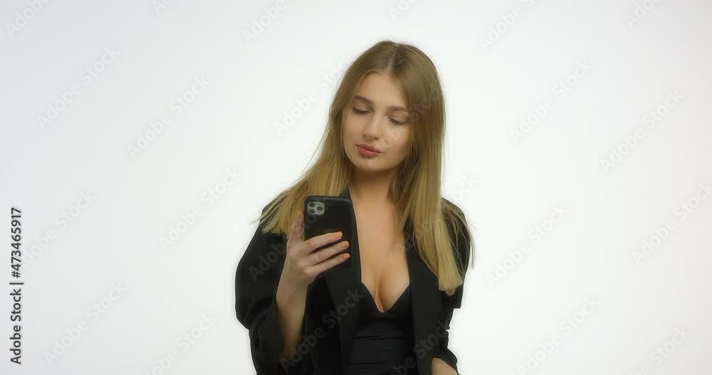 Beautiful woman with long blond hair in a black jacket is typing a message on the phone and is chatting. Girl model smiling and posing in the studio at the camera in the studio on a white background. Beautiful woman with long blond hair in a black jacket is typing a message on the phone and is chatting. Girl model smiling and posing in the studio at the camera in the studio on a white background.