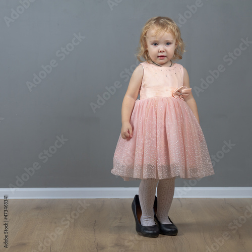 Cute baby girl 2 years old caucasian walks in black mom's big heels in pink prom dress